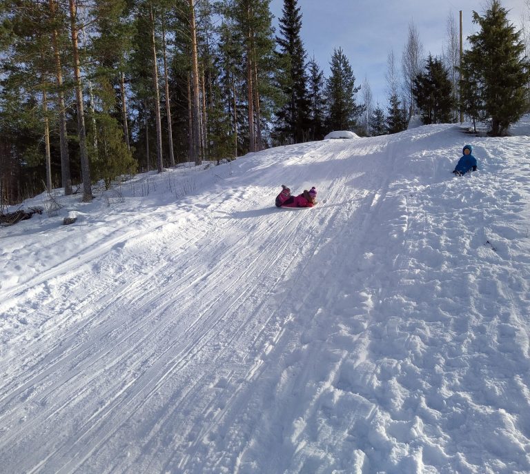 Laskiainen. On lunta. Lapsi laskee mäkeä liukurilla. Paljon pienempi lapsi istuu lumessa katsellen ihmeissään mäenlaskua.