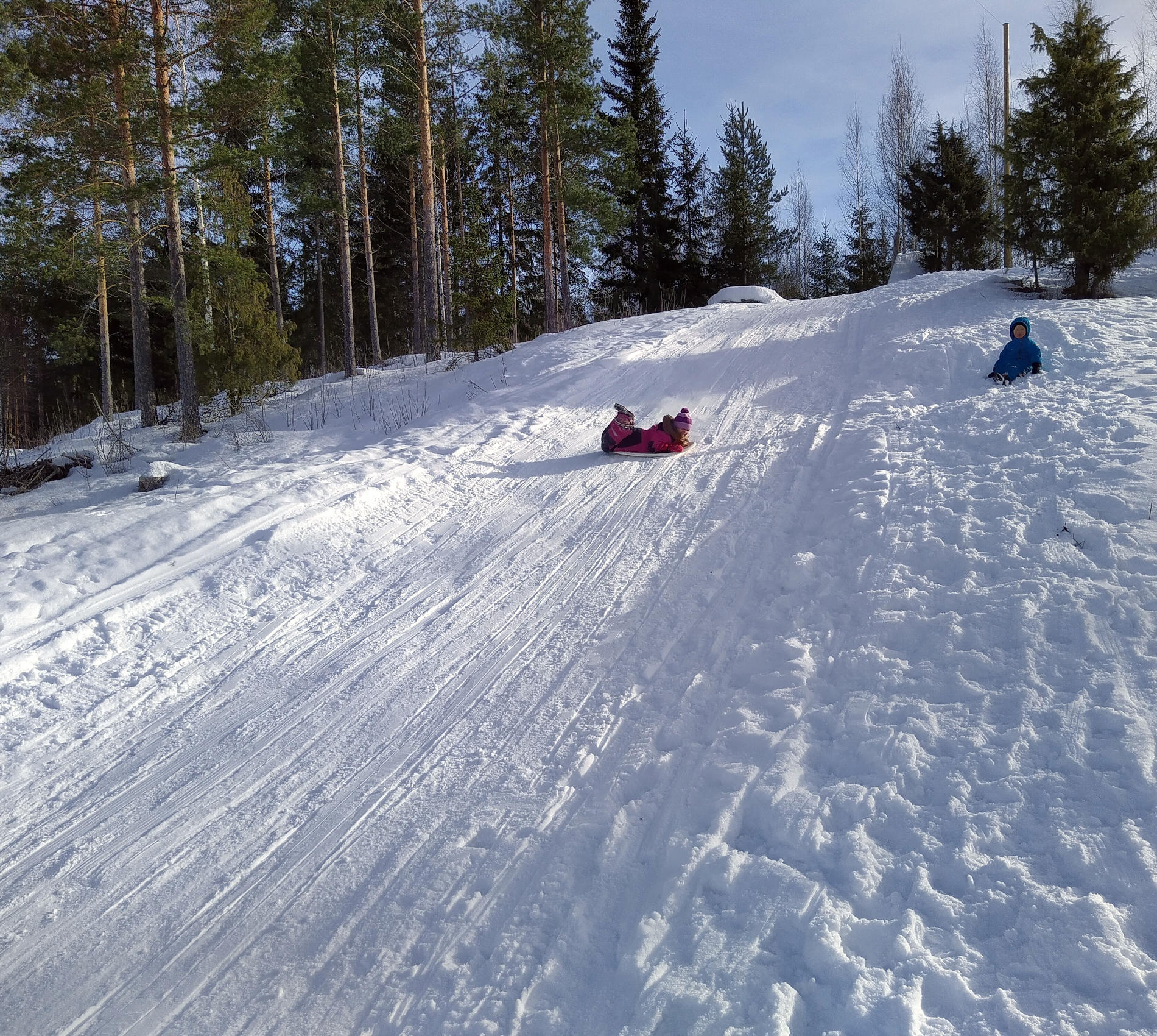 Laskiainen. On lunta. Lapsi laskee mäkeä liukurilla. Paljon pienempi lapsi istuu lumessa katsellen ihmeissään mäenlaskua.