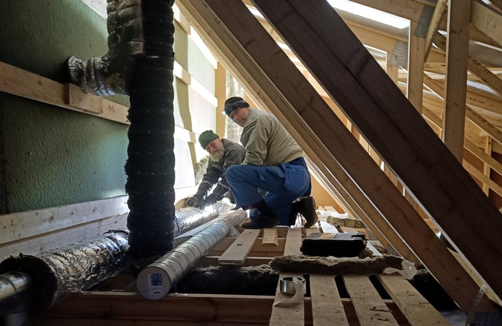 Kaksi rakennustyömiestä on ahtaassa paikassa kylmällä vintillä eristämässä iv-putkia